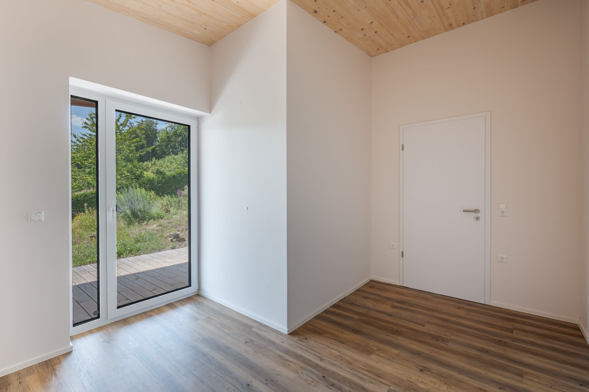 Leerer Raum mit Holzboden, weißen Wänden, Tür und großer Glas-Schiebetür mit Blick ins Grüne.