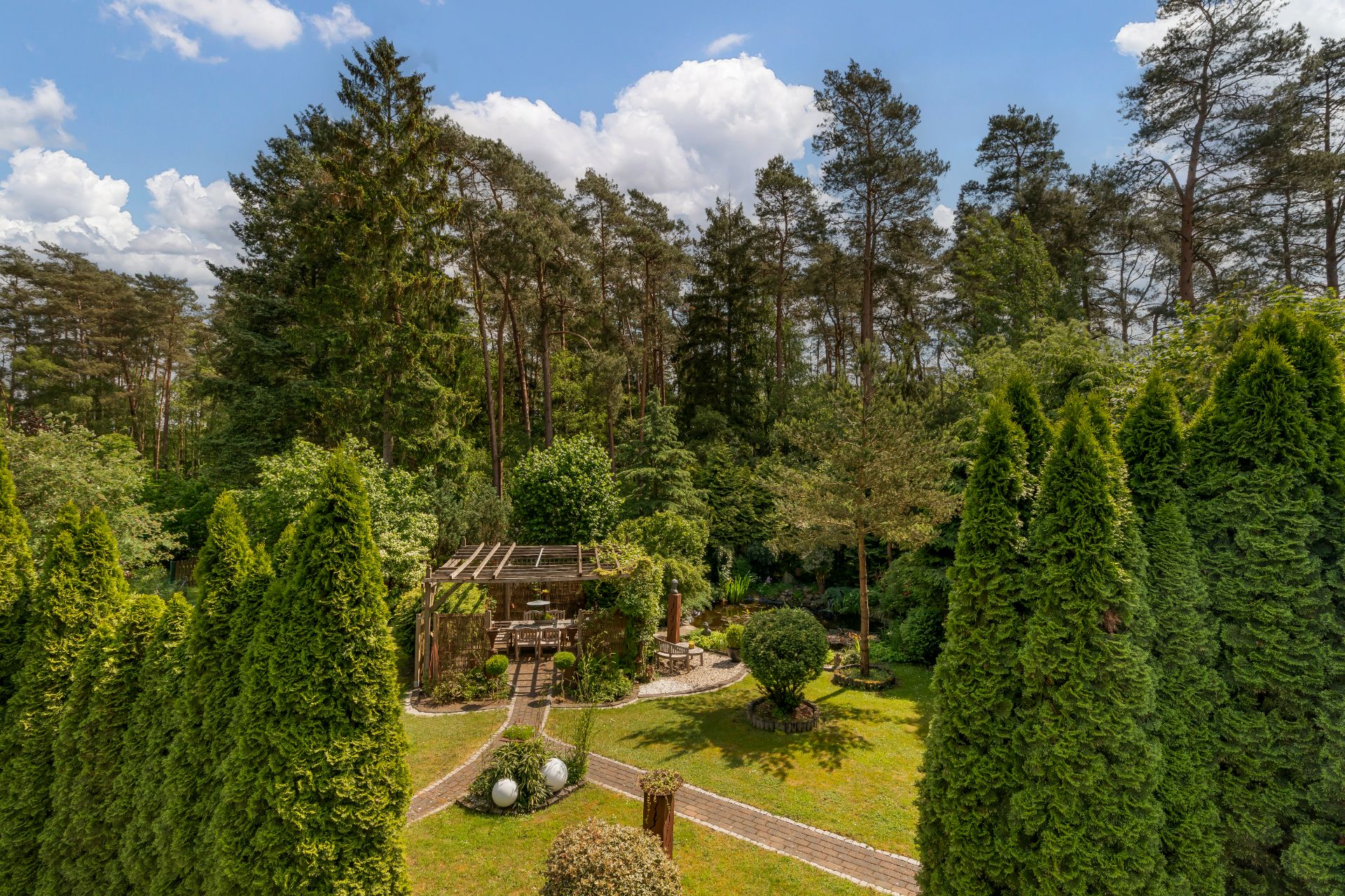 Gepflegter Garten mit Terrasse, Wegen und hohen Bäumen, im Hintergrund dichter Wald.