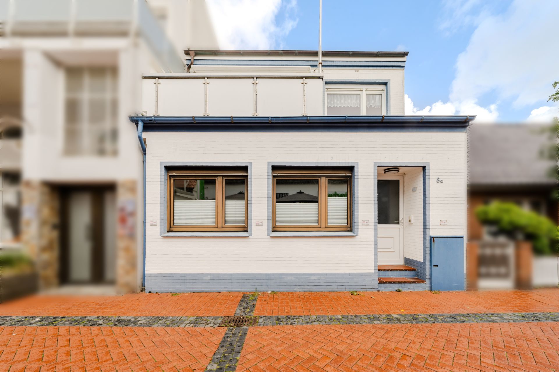 Weiße Wohnhausfassade mit zwei Fenstern und einer Haustür, davor gepflasterter Vorplatz.