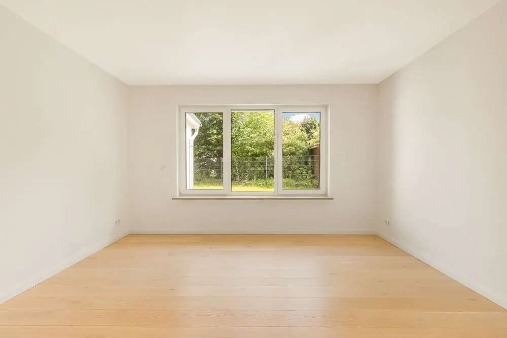 Leerer Raum mit hellem Holzfußboden und großem Fenster mit Blick auf einen Garten.
