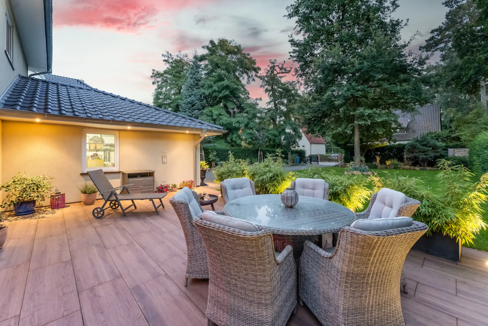 Große Terrasse mit rundem Tisch, sechs Stühlen, Liege und Pflanzen, Blick auf Garten bei Sonnenuntergang.