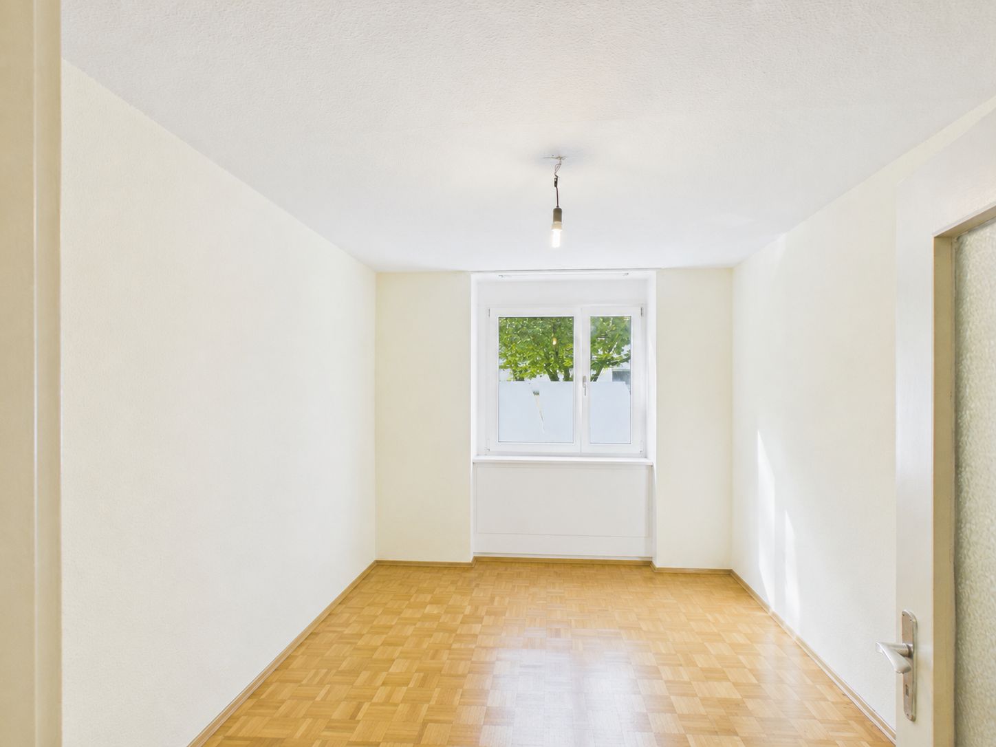 Heller leerer Raum mit Parkettboden, weißem Fenster und Glühbirne an der Decke.