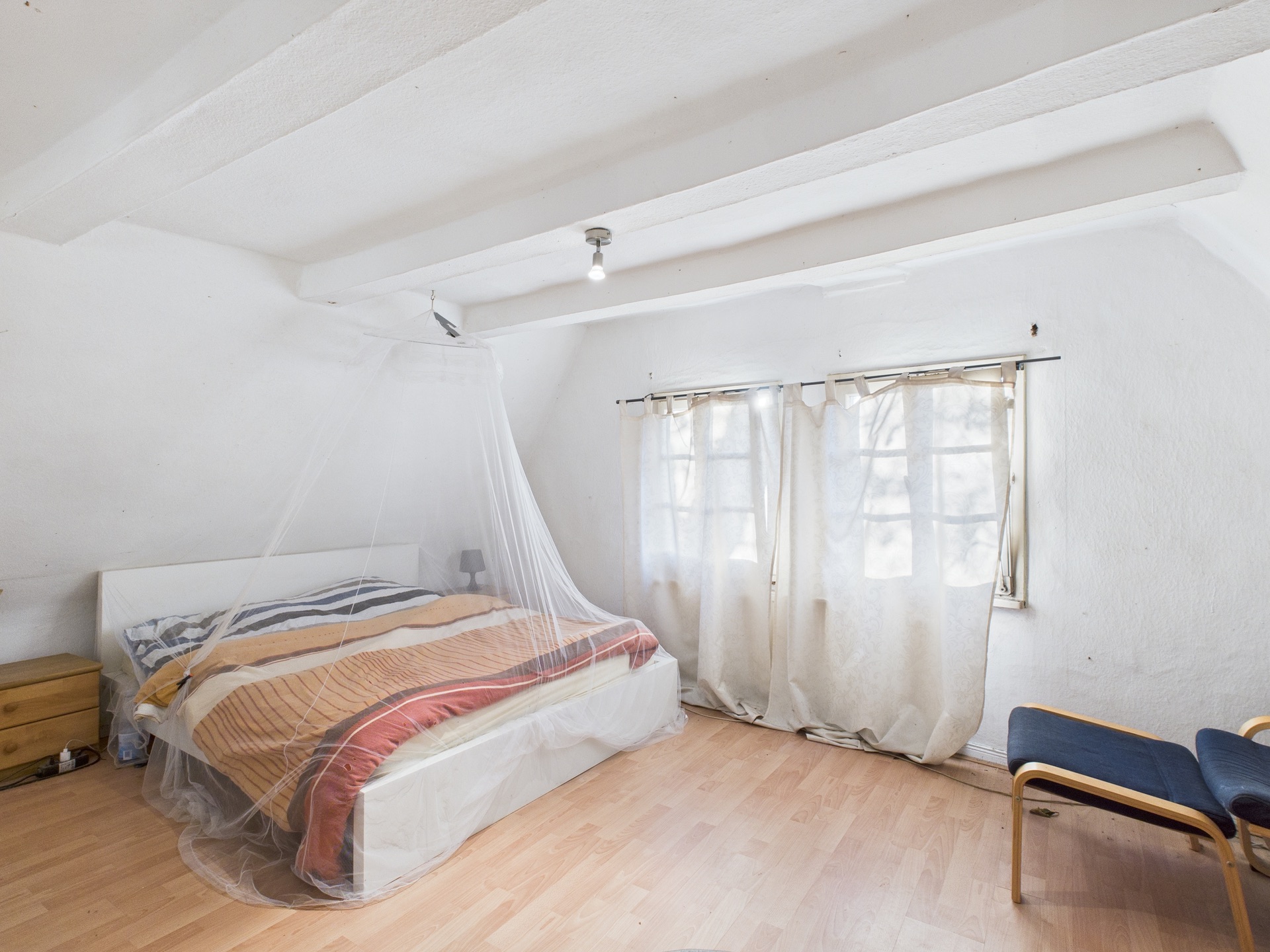 Schlafzimmer mit Doppelbett, Moskitonetz, zwei Fenstern mit Vorhängen und Holzfußboden.