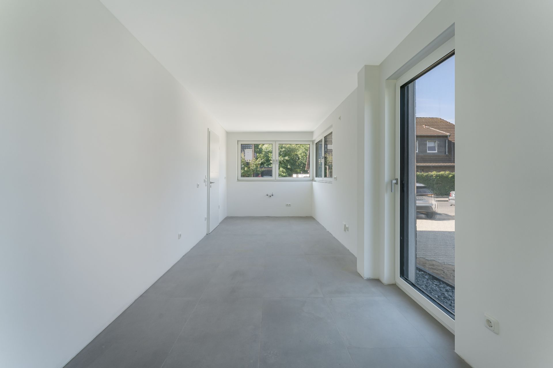 Heller, leerer Raum mit großen Fenstern und grauem Fliesenboden in einem Wohnhaus.