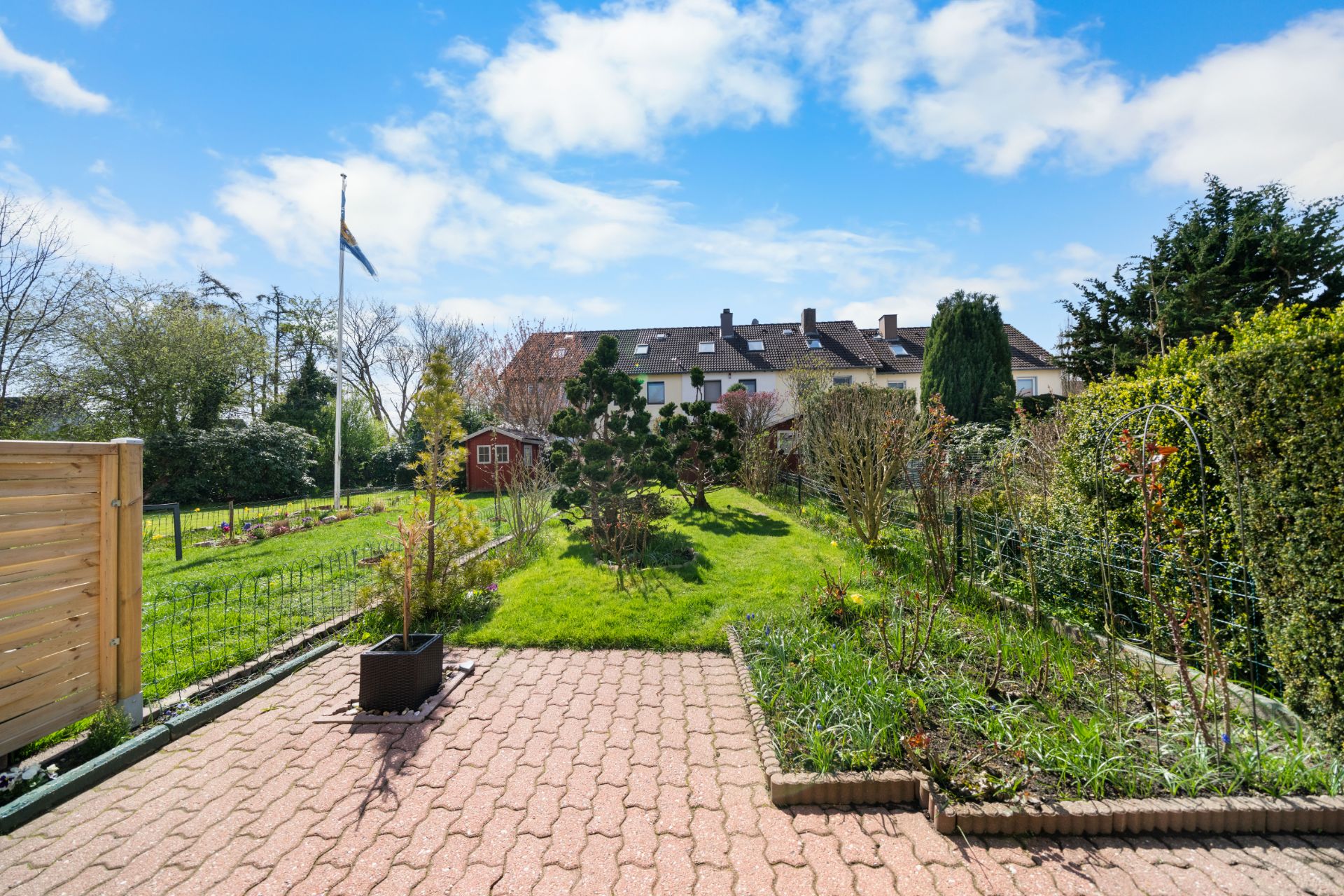 Gepflasterte Terrasse mit Blick auf Garten, Rasenfläche und Sträucher, im Hintergrund Reihenhäuser.