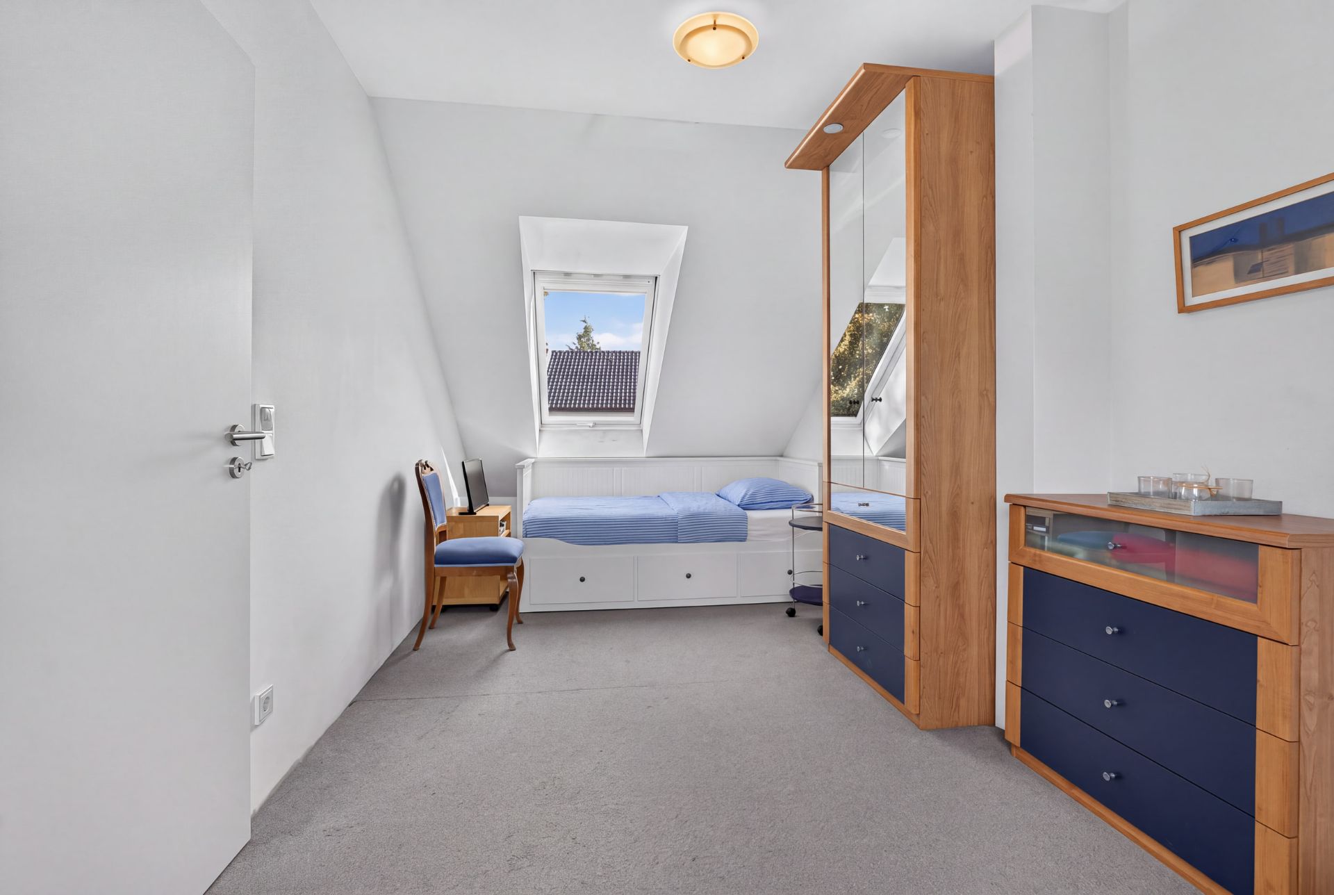 Helles Schlafzimmer mit Einzelbett, Schrank, Kommode und Stuhl unter Dachschräge.