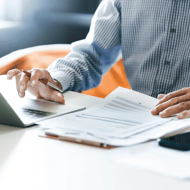 Person arbeitet am Laptop mit Dokumenten und Smartphone auf einem Tisch.