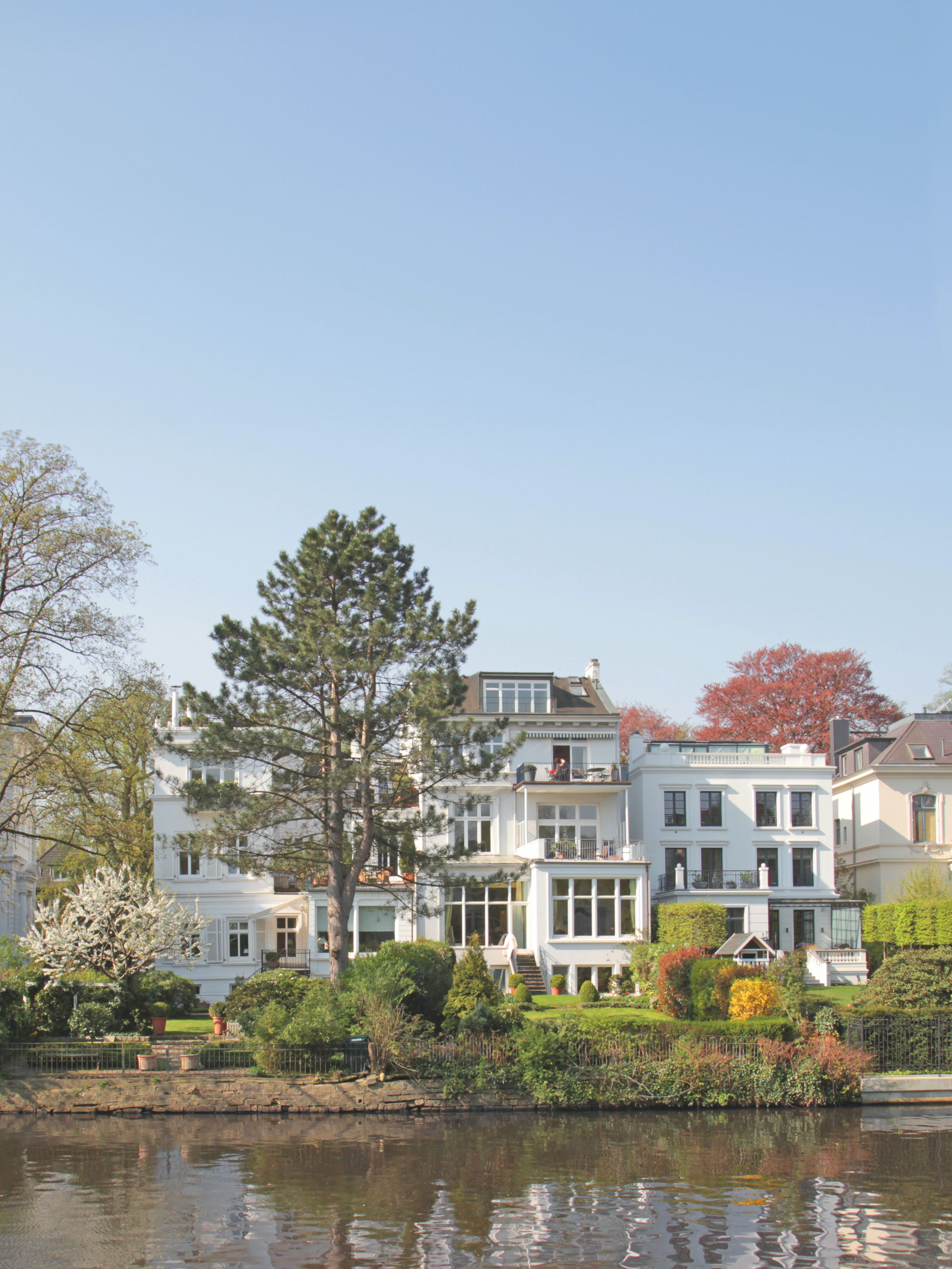 Großes weißes Wohnhaus mit Garten am Wasser, Bäume und Büsche im Vordergrund.