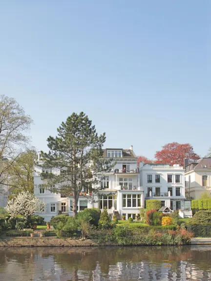 Großes weißes Wohnhaus mit Garten am Wasser, Bäume und Büsche im Vordergrund.