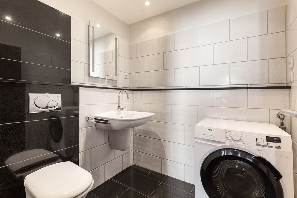 Modernes Bad mit WC, Wandwaschbecken, Spiegel und Waschmaschine, weiße Wandfliesen.
