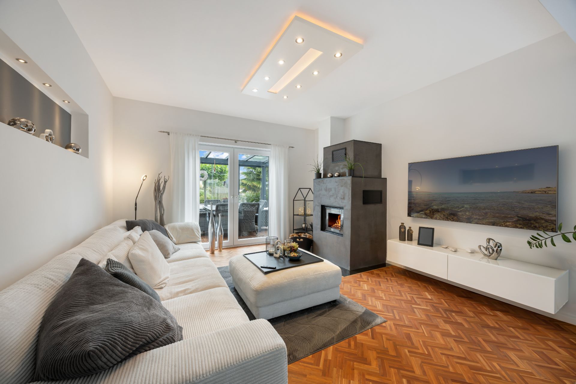 Modernes Wohnzimmer mit weißem Sofa, Kamin, großem Fernseher und Zugang zur Terrasse.