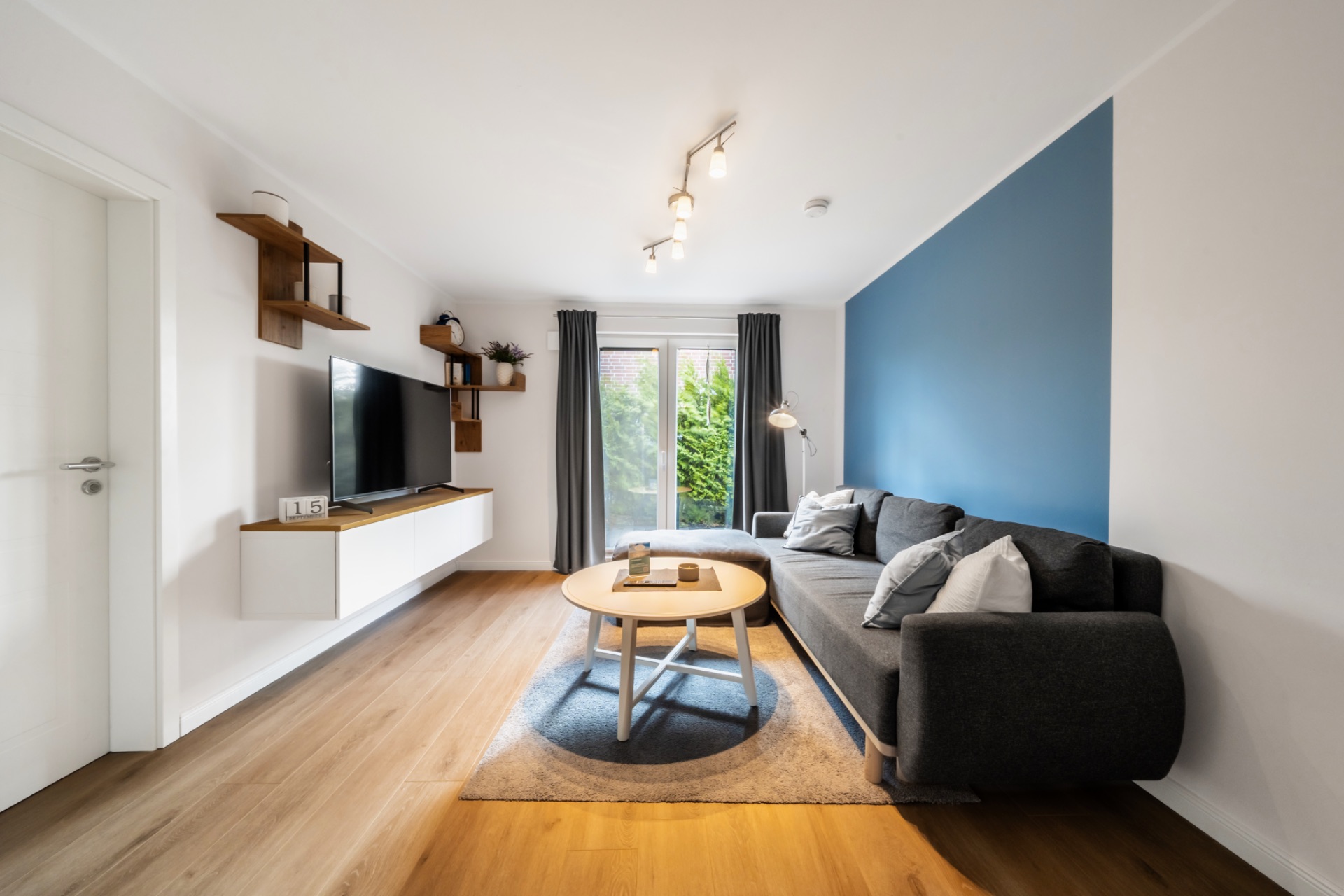 Modernes Wohnzimmer mit grauem Sofa, rundem Tisch, Fernseher und großer Fensterfront mit Vorhängen.