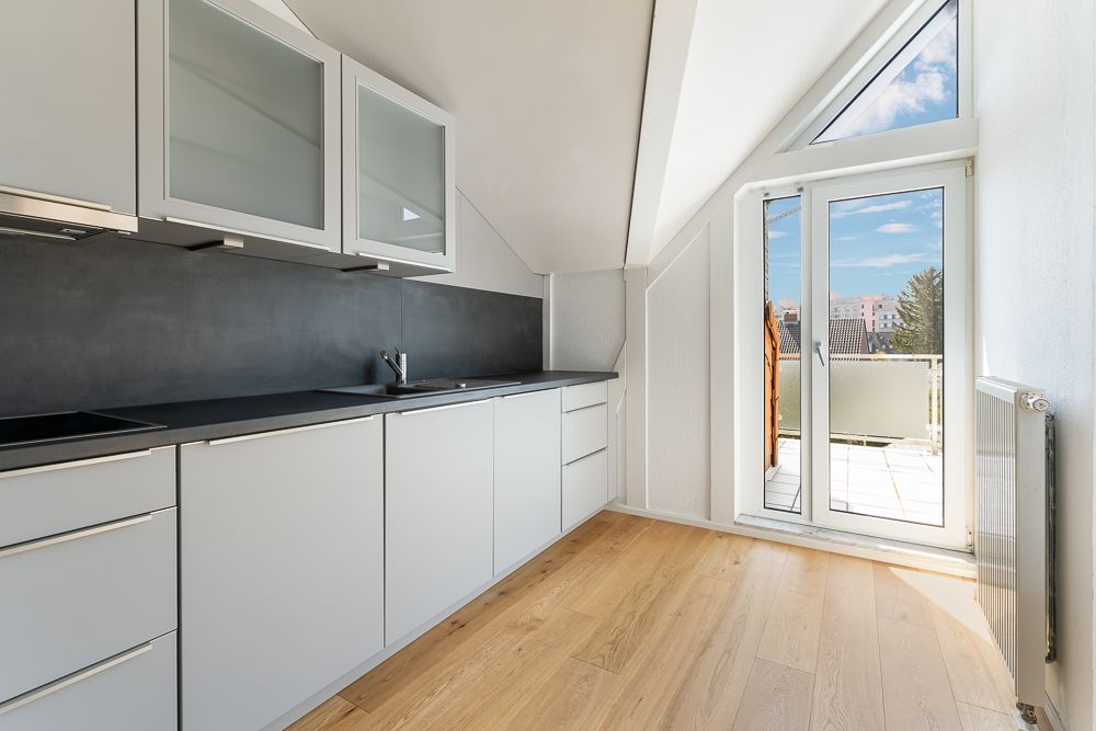 Moderne Küche mit hellen Schränken, Holzboden und Zugang zu einem Balkon mit Glas­türen.