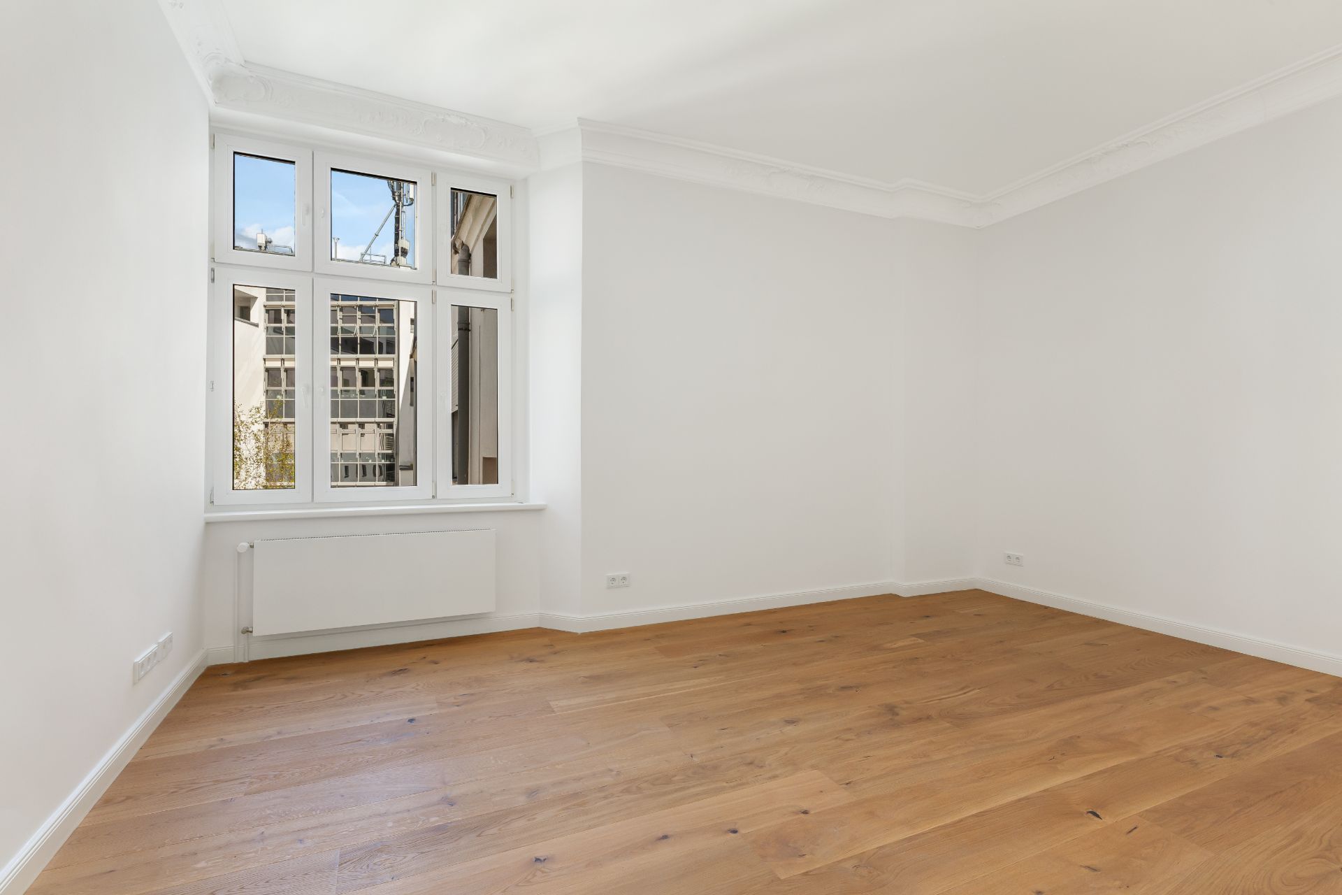 Leerer, heller Raum mit großem Fenster, weißen Wänden und Holzfußboden.