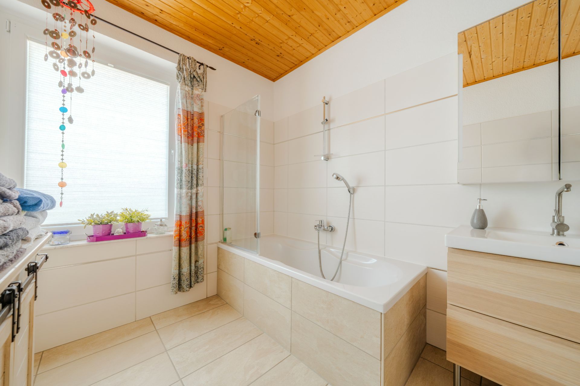 Helles Badezimmer mit Badewanne, Waschbecken, Spiegel und Fenster mit Vorhang.