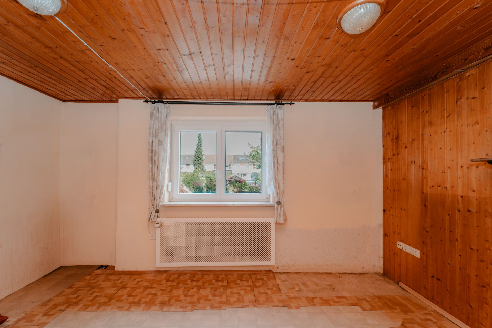 Leeres Zimmer mit Holzdecke, Holzwand, Fenster mit Vorhängen und sichtbarem Heizkörper.