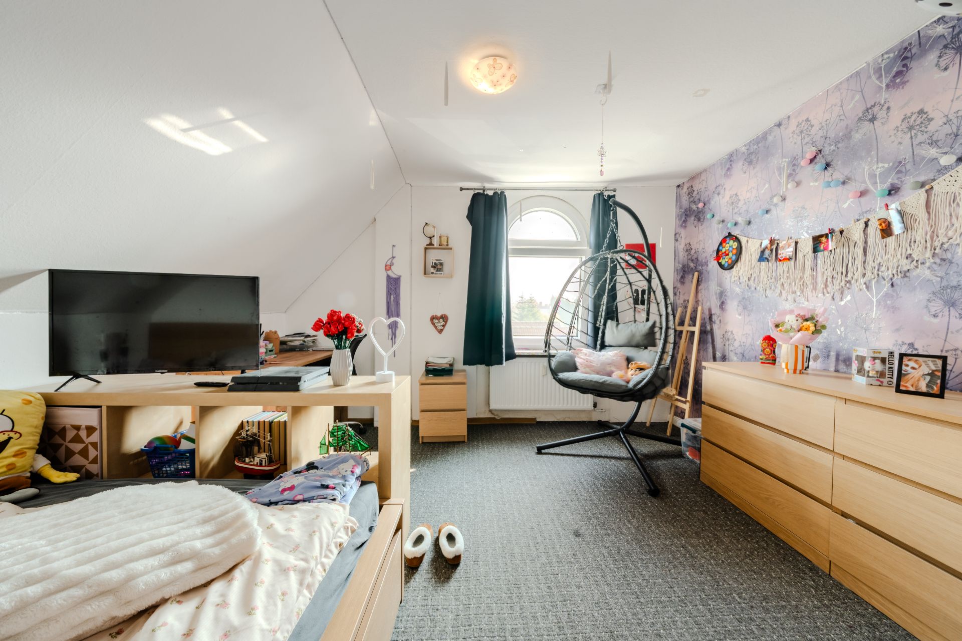 Helles Schlafzimmer mit Bett, Fernseher, Kommode und Hängesessel vor einem Fenster.
