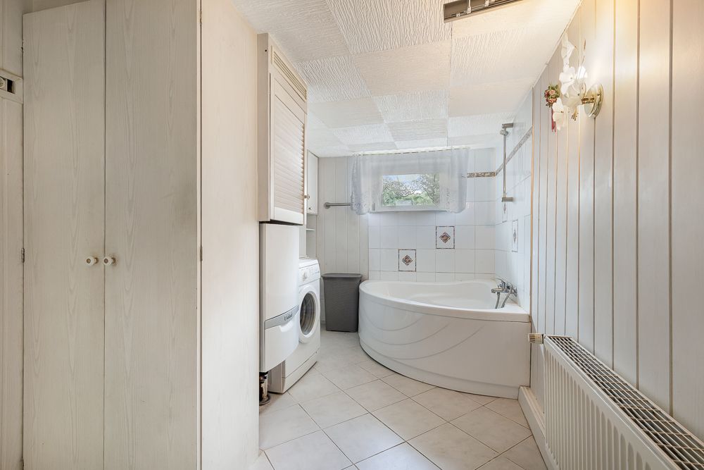 Helles Badezimmer mit Eckbadewanne, Waschmaschine, Schrank und kleinem Fenster.