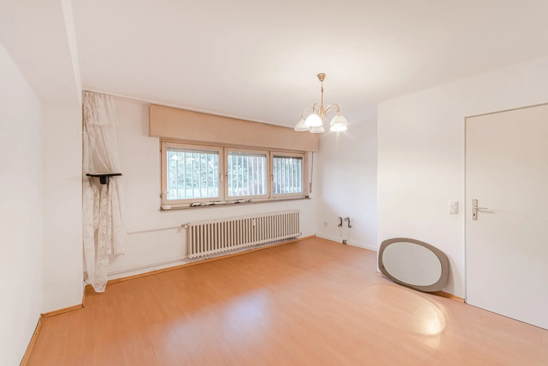 Leerer Raum mit Holzfußboden, drei Fenstern mit Gitter, Heizkörper und Deckenlampe.