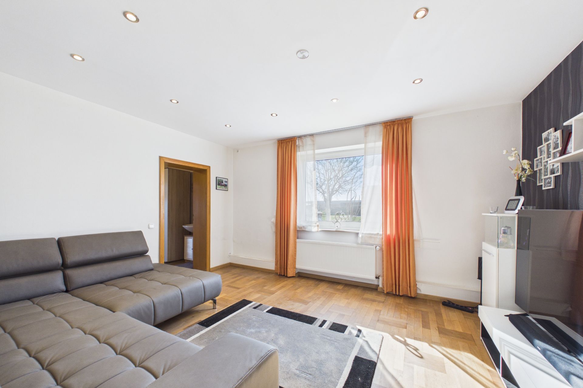 Helles Wohnzimmer mit grauem Sofa, Fenster mit orangefarbenen Vorhängen und Parkettboden.