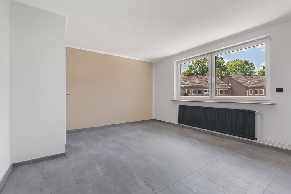 Heller, leerer Raum mit großen Fenstern, grauen Fliesen und beige gestrichener Wand.