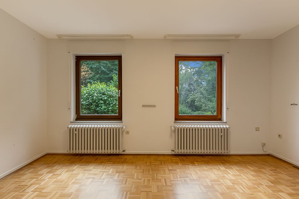 Leeres Zimmer mit Parkettboden, zwei Fenstern und zwei Heizkörpern unter den Fenstern.