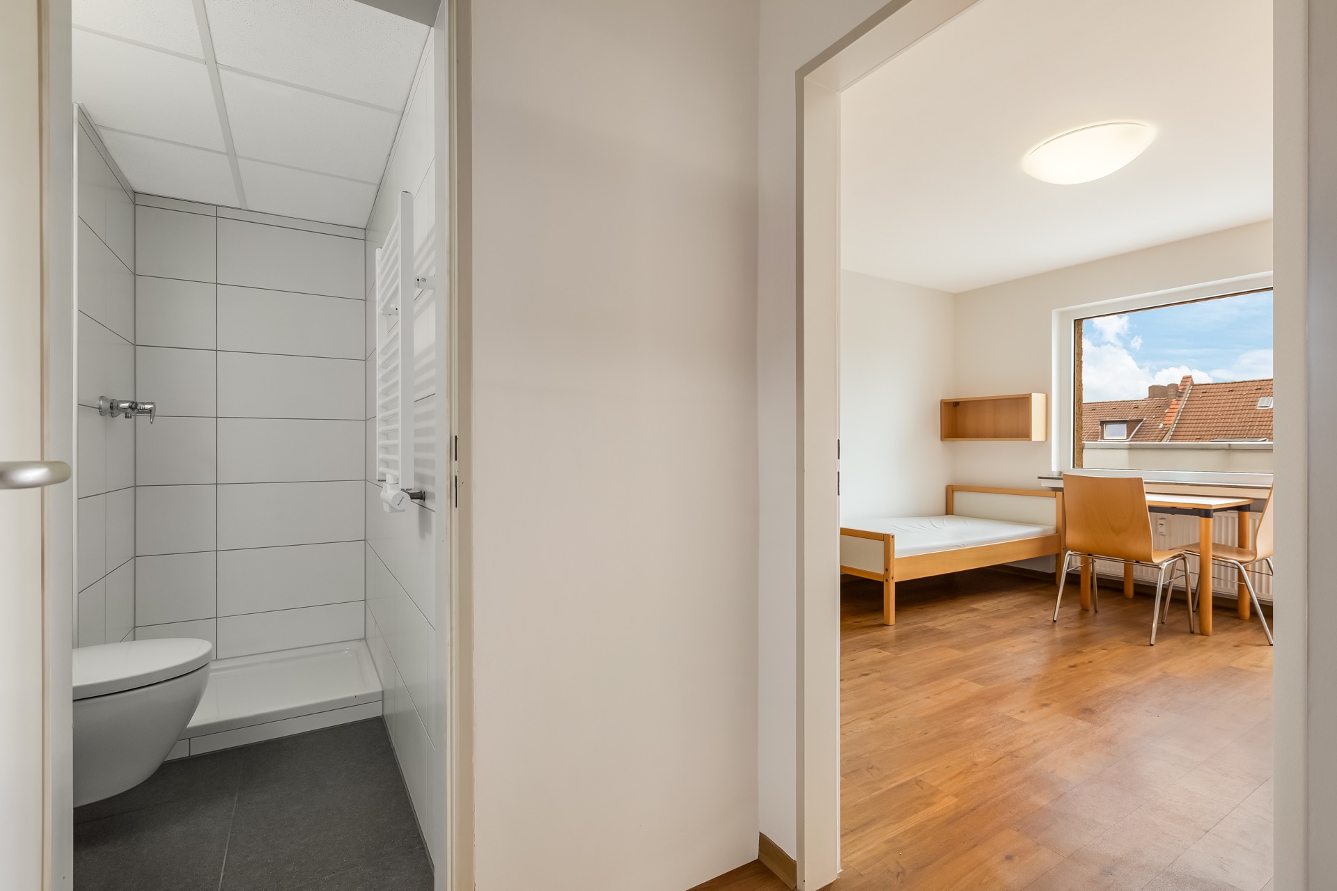 Blick vom Flur auf ein kleines Bad und ein helles Zimmer mit Bett, Tisch und Stühlen.