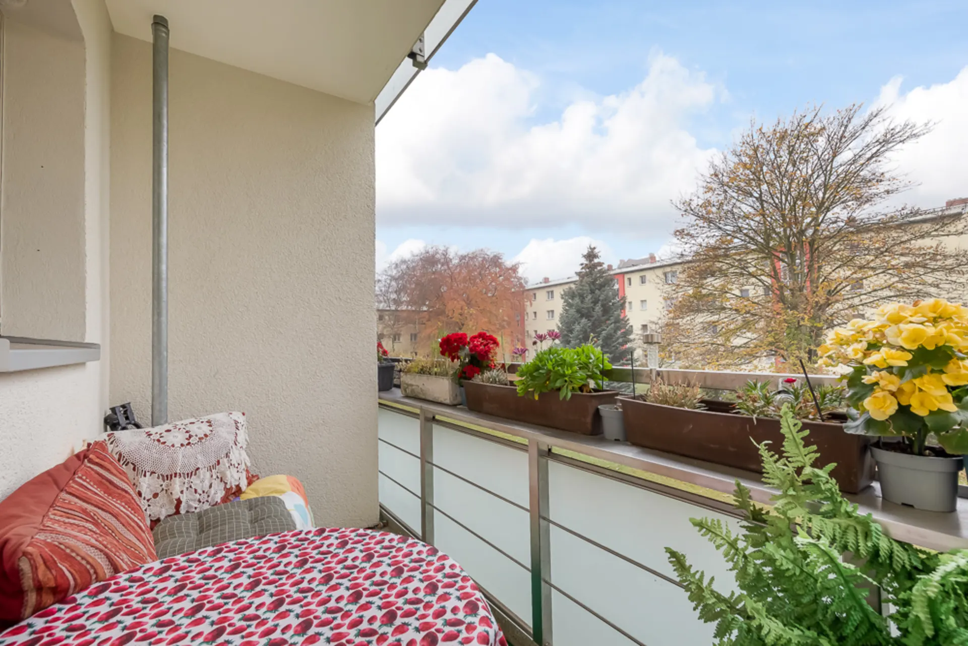 Balkon mit Blumen und Pflanzen, ein Stuhl mit Kissen und Blick auf Bäume und Wohngebäude.
