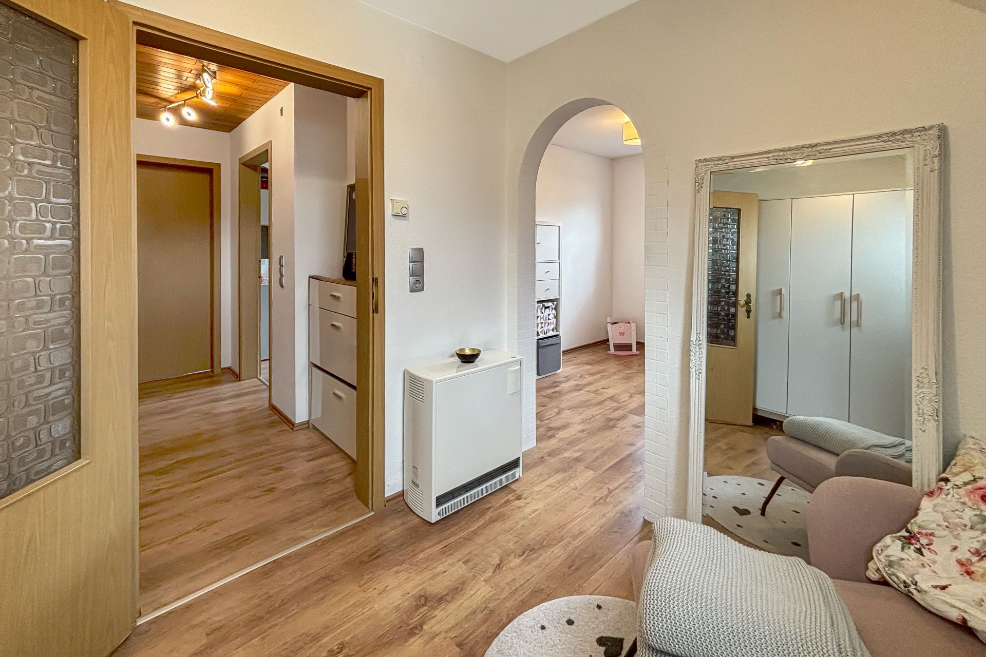 Wohnungsflur mit Holzfußboden, offenem Durchgangsbogen und Spiegel, angrenzendes Zimmer sichtbar.