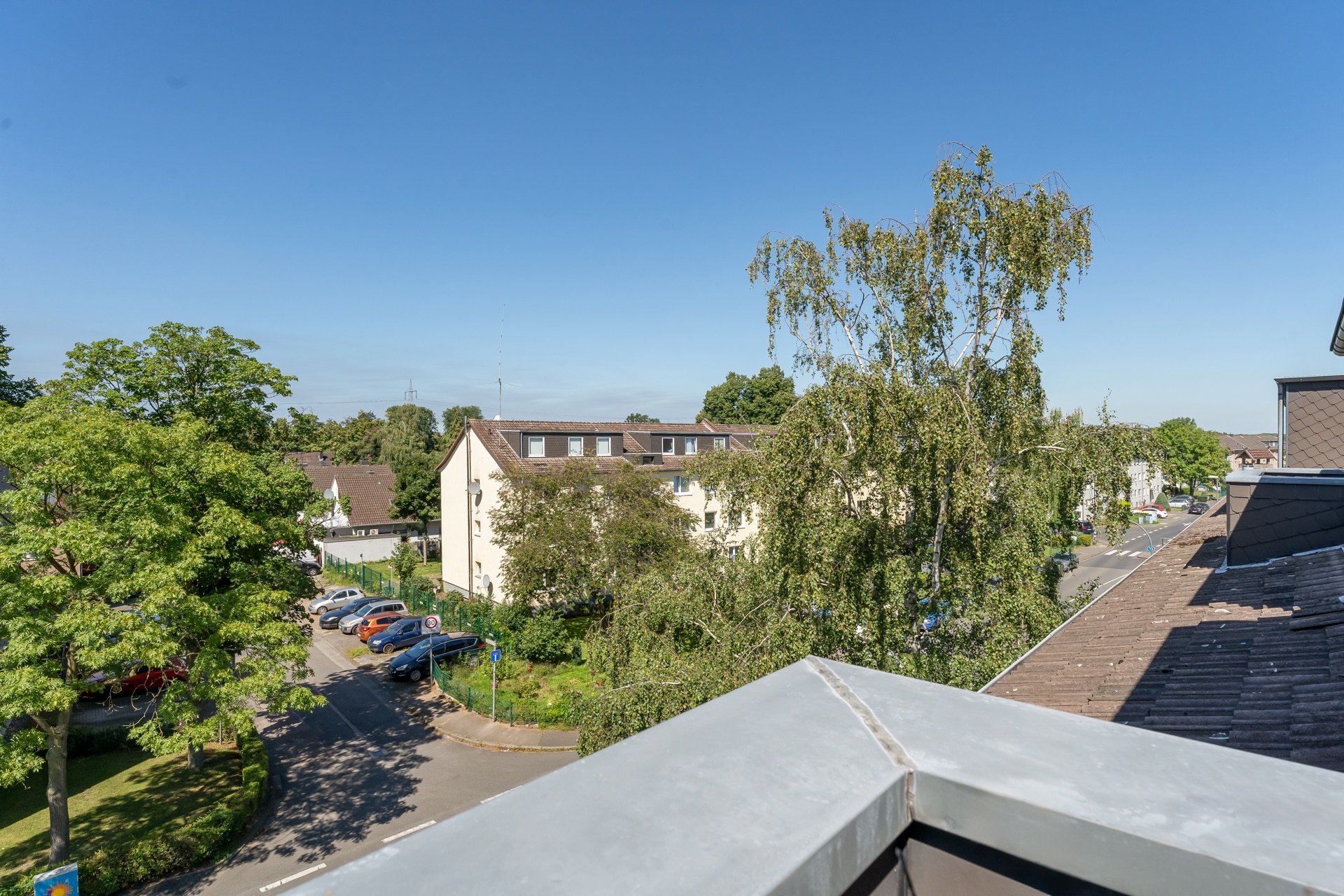 Blick von einem Balkon auf Wohnhäuser, Bäume und eine Straße mit geparkten Autos.