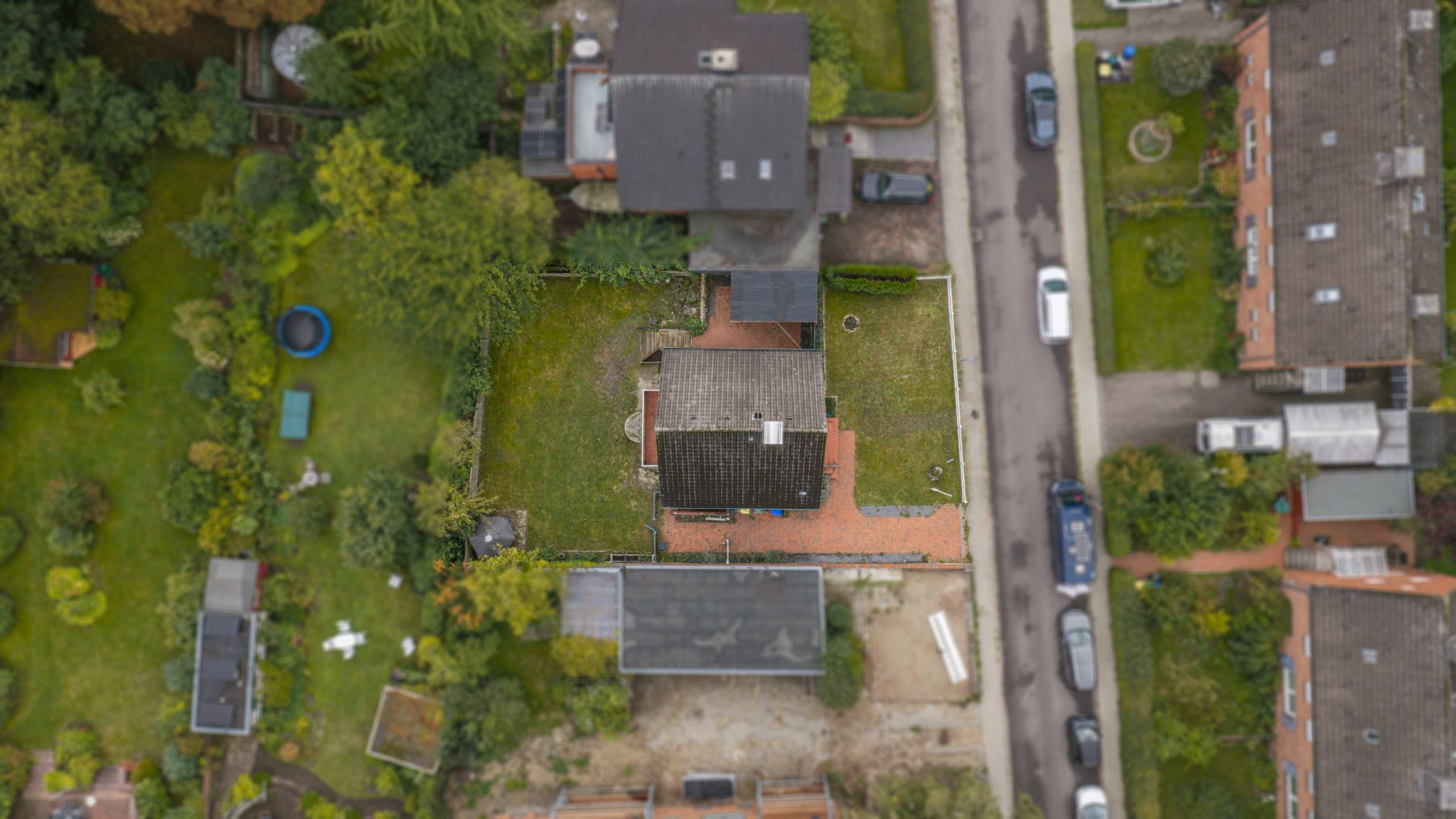 Luftaufnahme eines Hauses mit Garten, umgeben von weiteren Häusern und einer Straße mit parkenden Autos.