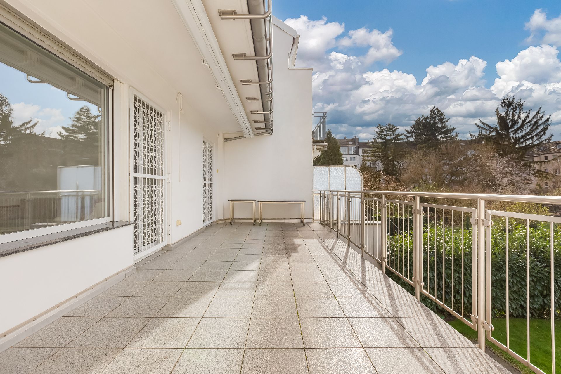 Großer Balkon mit Fliesenboden und Metallgeländer, Blick auf Garten und Bäume.