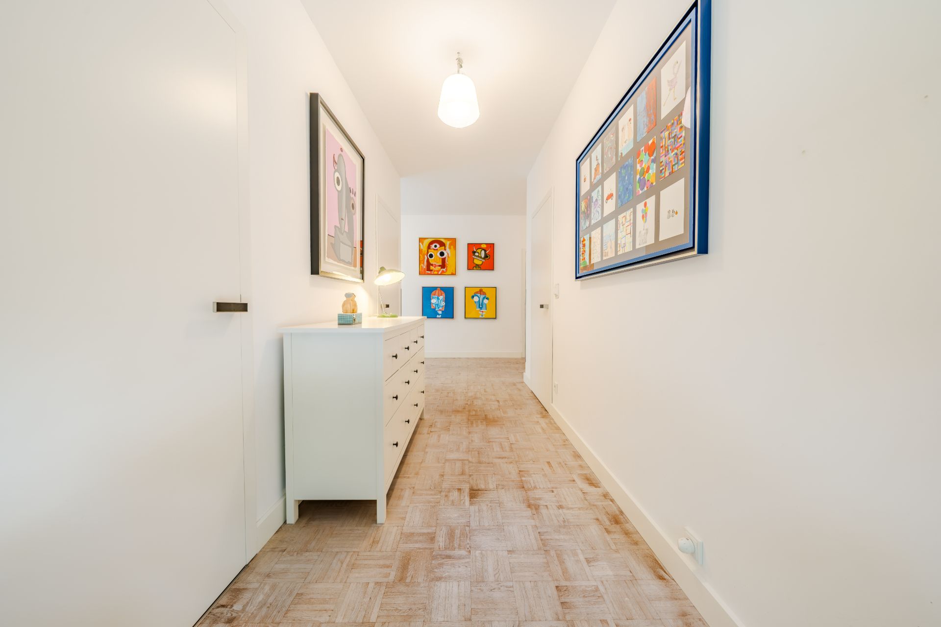 Heller Flur mit weißer Kommode, Holzboden und bunten Bildern an den Wänden.
