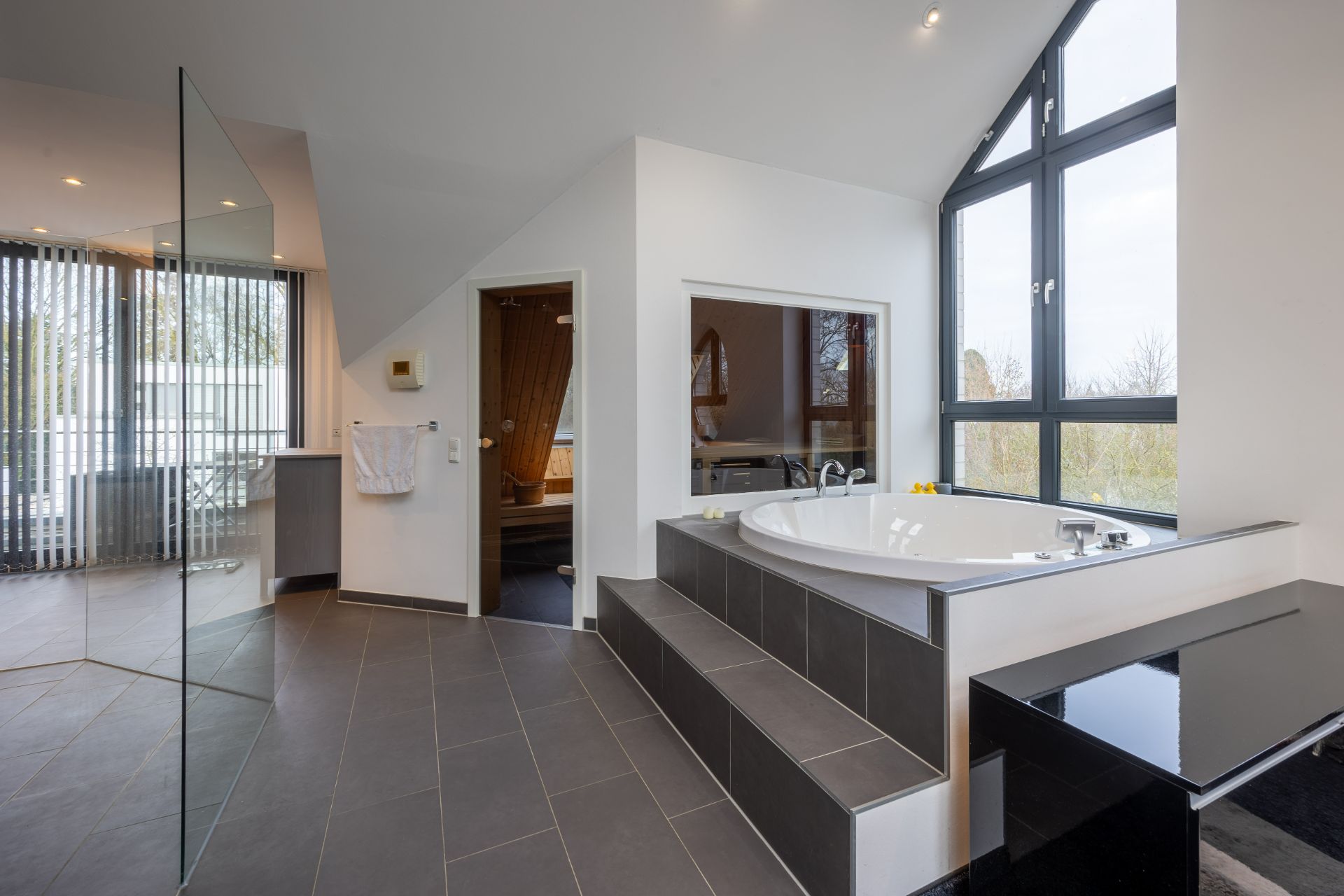 Modernes Badezimmer mit großer Badewanne, Glasdusche, Fenstern und Zugang zur Sauna.