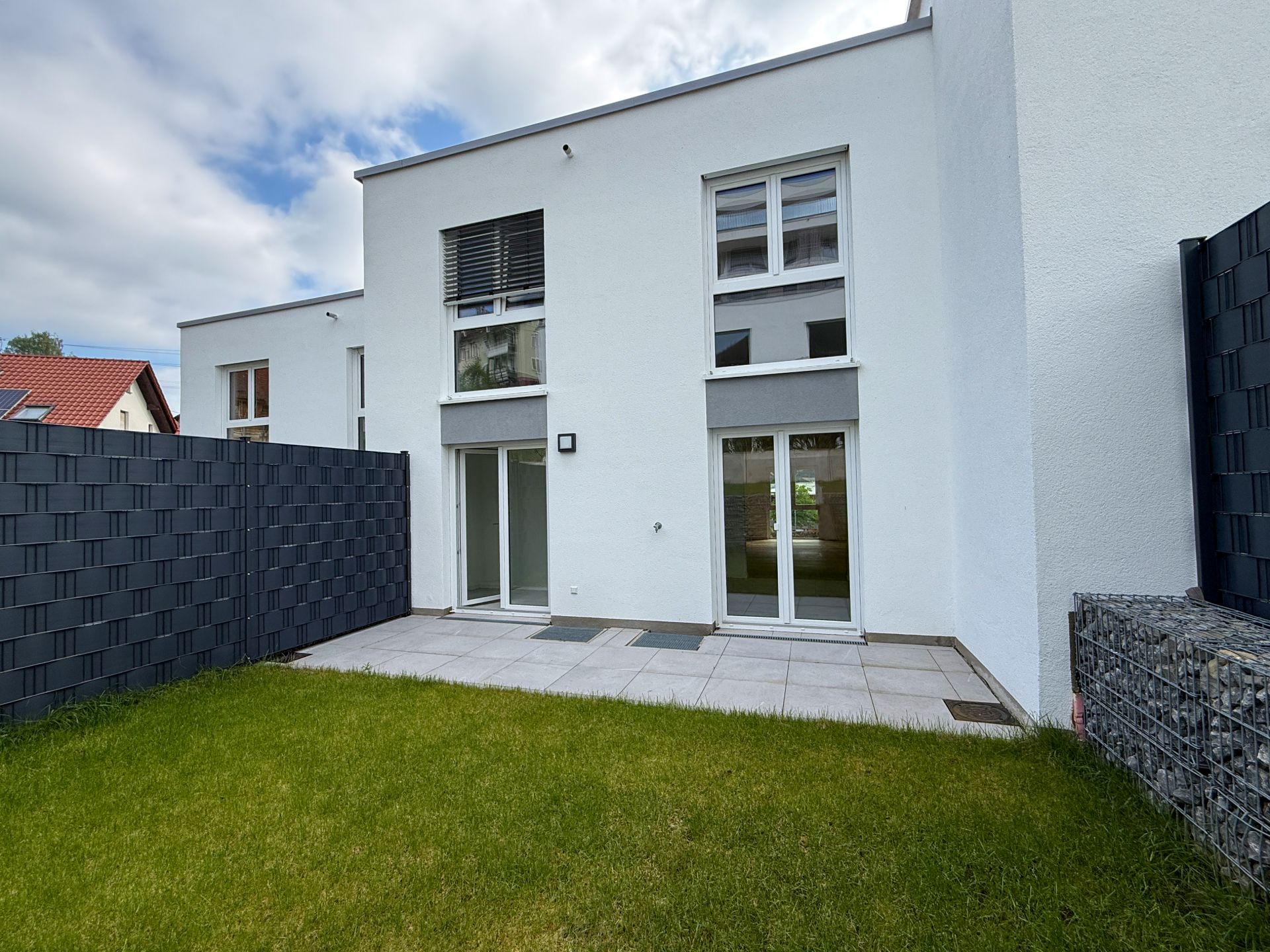 Moderne weiße Hausfassade mit Terrasse und kleinem Garten, seitlich von Zäunen eingerahmt.