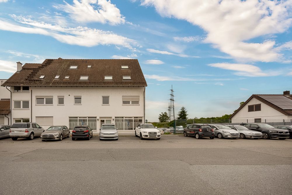 Mehrstöckiges Wohngebäude mit schrägem Dach und parkenden Autos davor bei blauem Himmel.