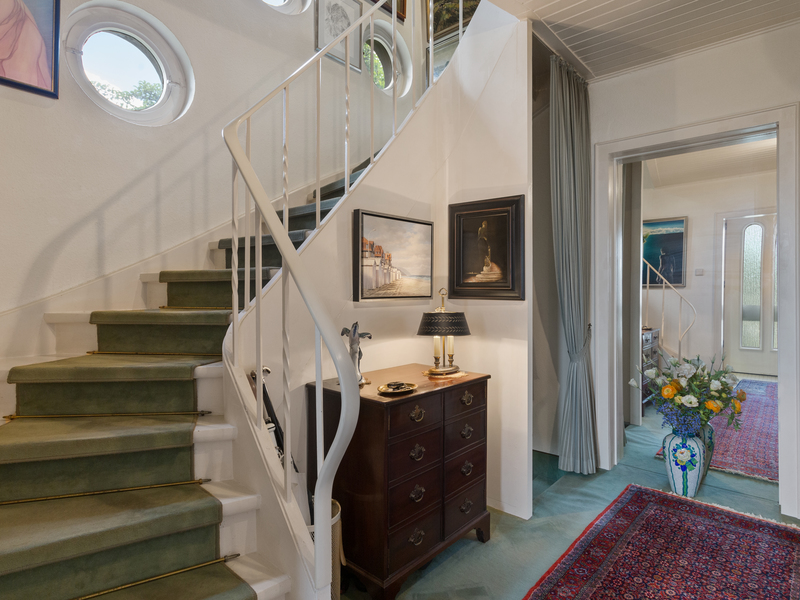 Heller Flur mit grüner Treppe, Kommode, Tischlampe, Blumenstrauß und runden Fenstern.