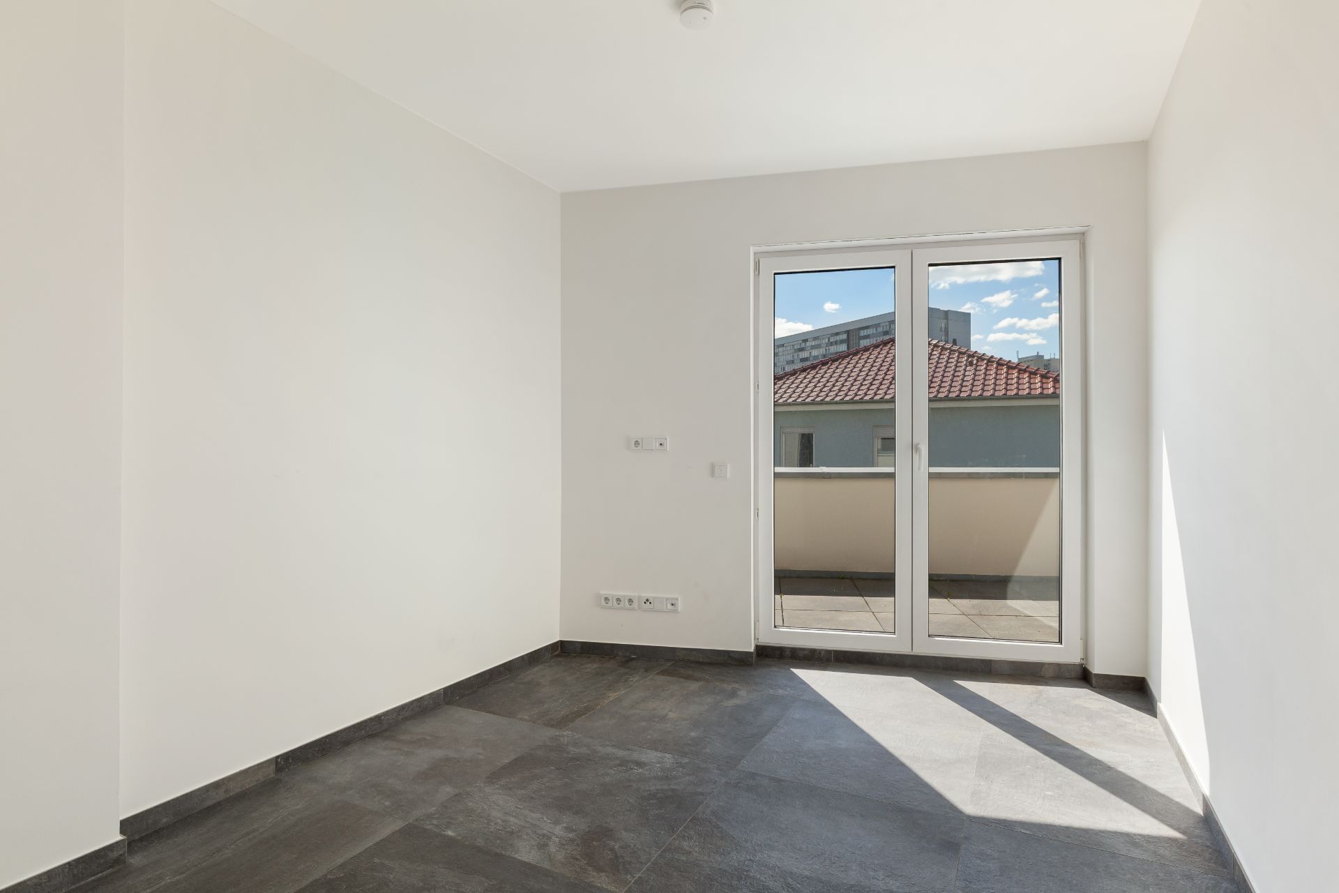 Leerer, heller Raum mit dunklem Boden und Glastüren zu einem Balkon.