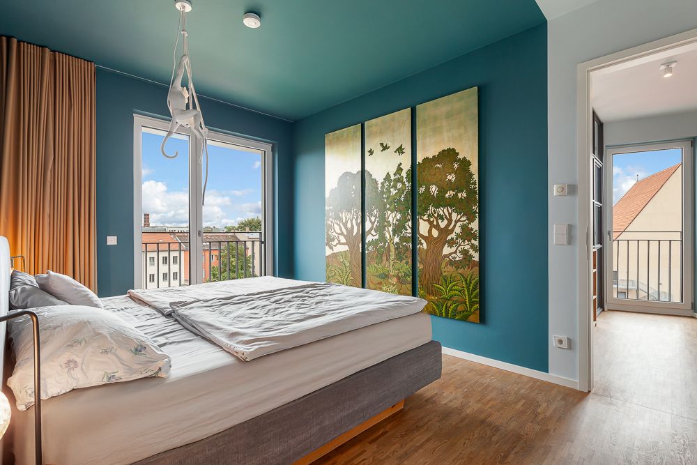 Modernes Schlafzimmer mit großem Bett, Wandbild mit Bäumen und Zugang zu Balkon.