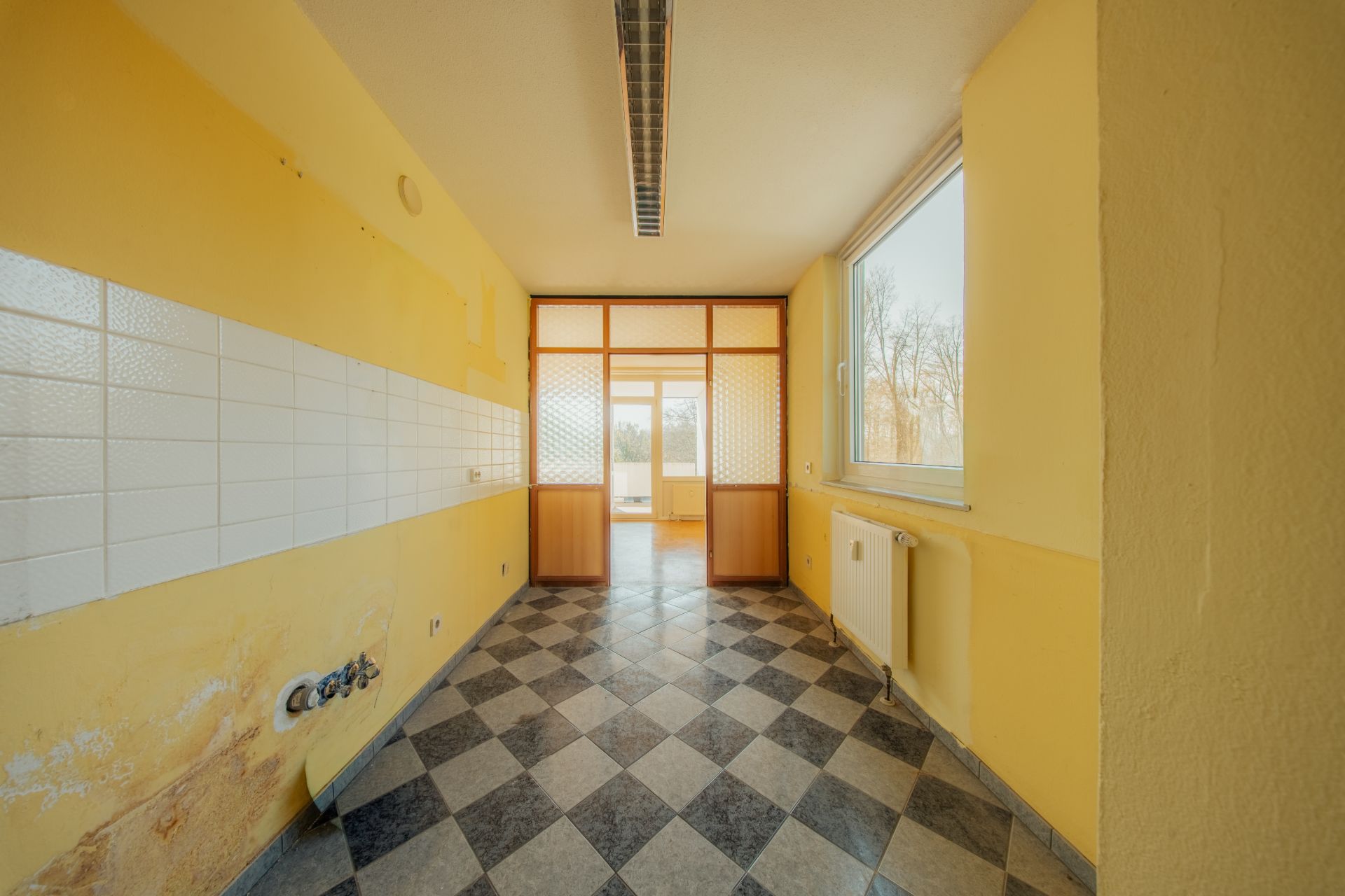 Leerer Raum mit gelben Wänden, Fliesenboden und großem Fenster, Türen führen zum nächsten Zimmer.