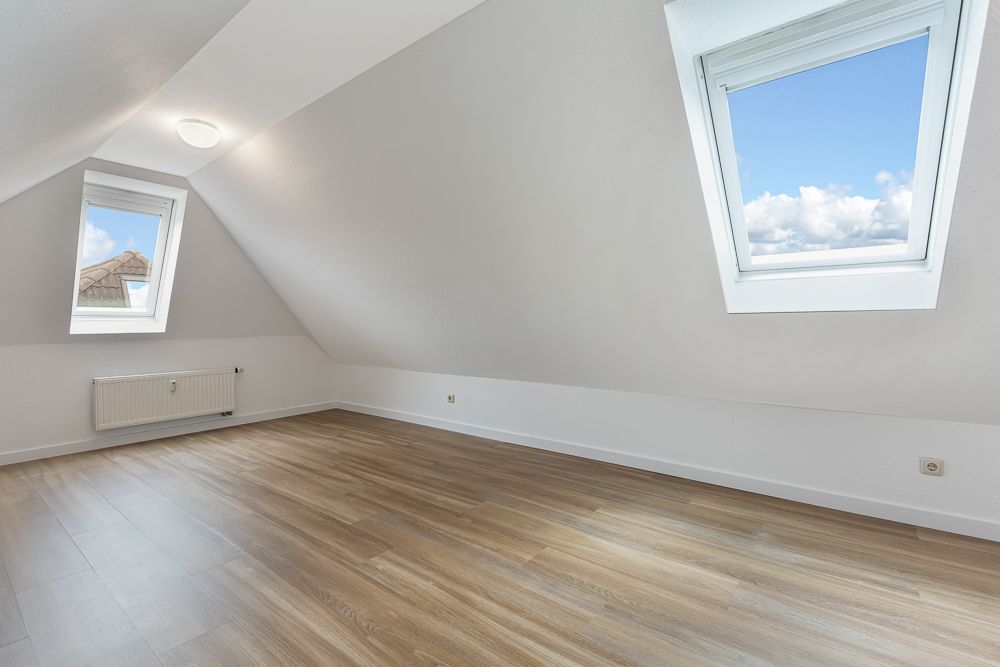 Leerer Dachgeschossraum mit schrägen Wänden, zwei Fenstern und hellem Holzboden.
