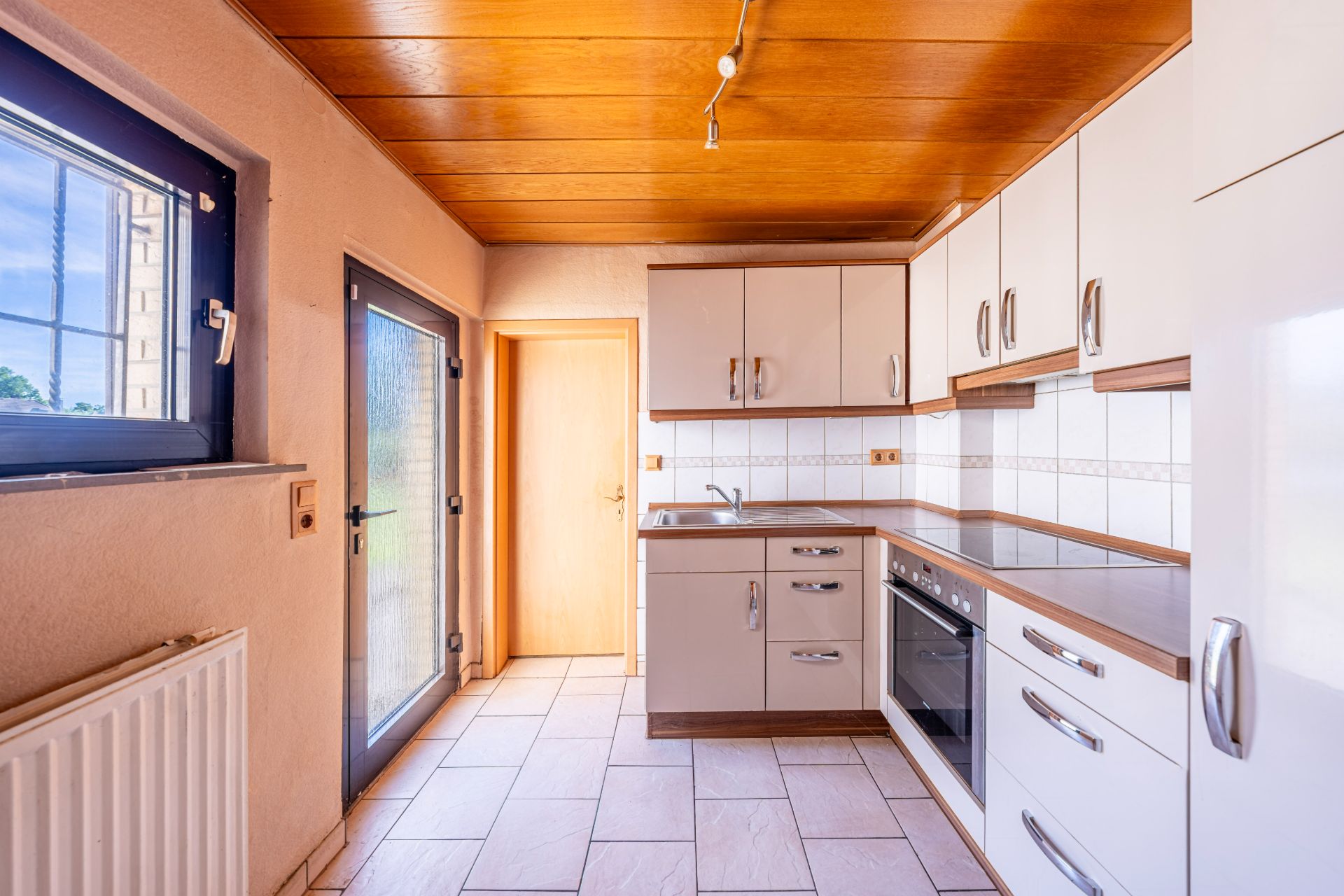 Helle Einbauküche mit weißen Schränken, Herd, Spüle und Fliesenboden, Tür und Fenster.