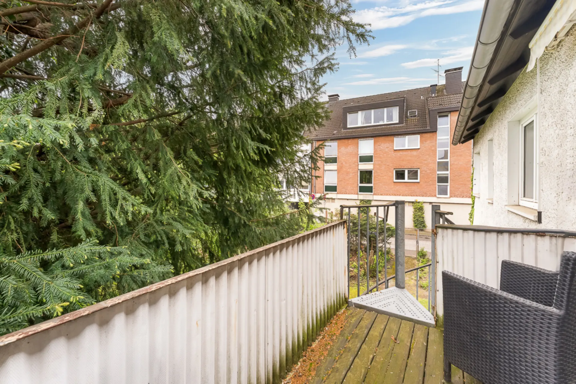 Kleiner Balkon mit Stuhl, Blick auf grünen Baum und Wohngebäude gegenüber.
