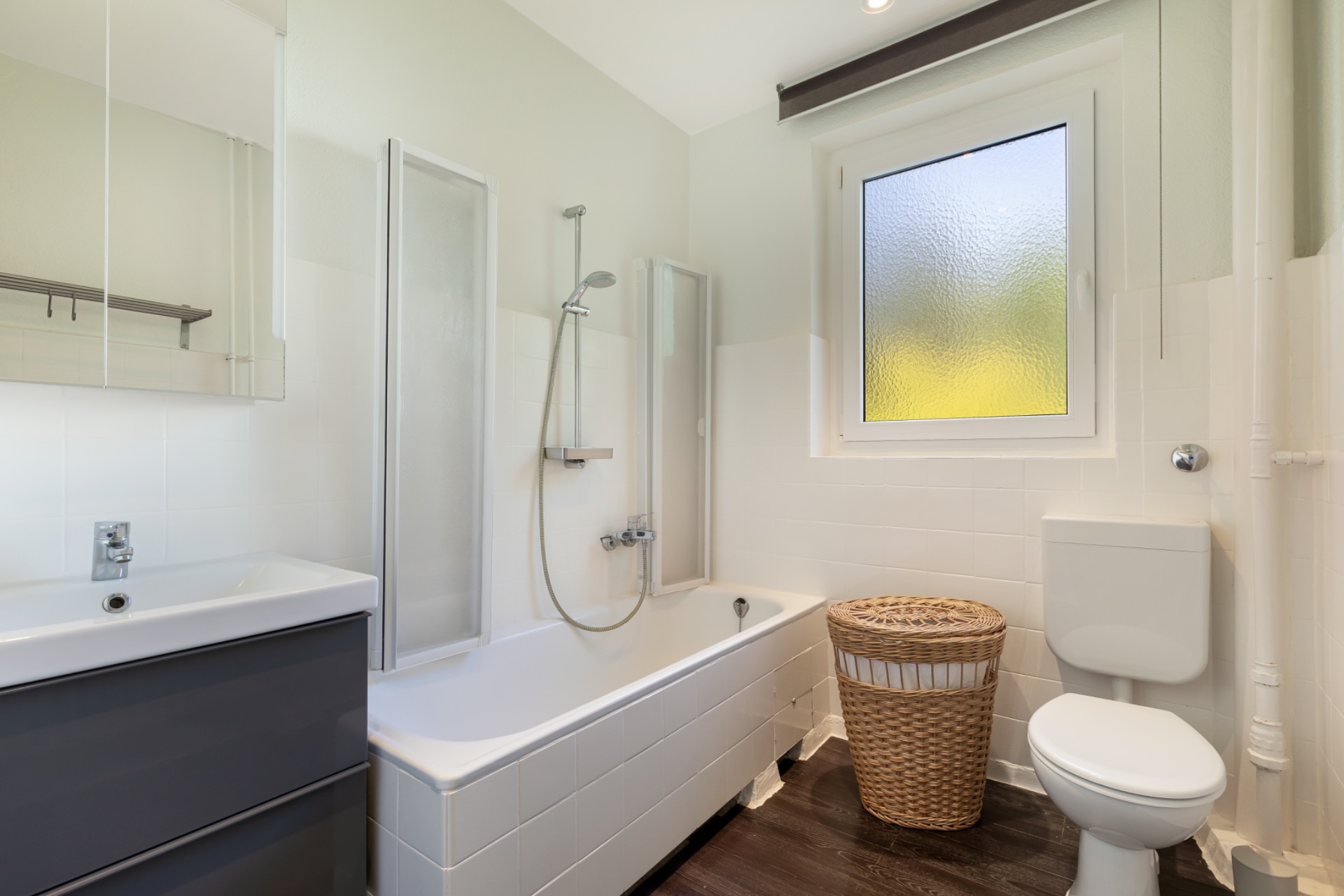 Helles Badezimmer mit Badewanne, Waschbecken, Toilette, Fenster und Wäschekorb.