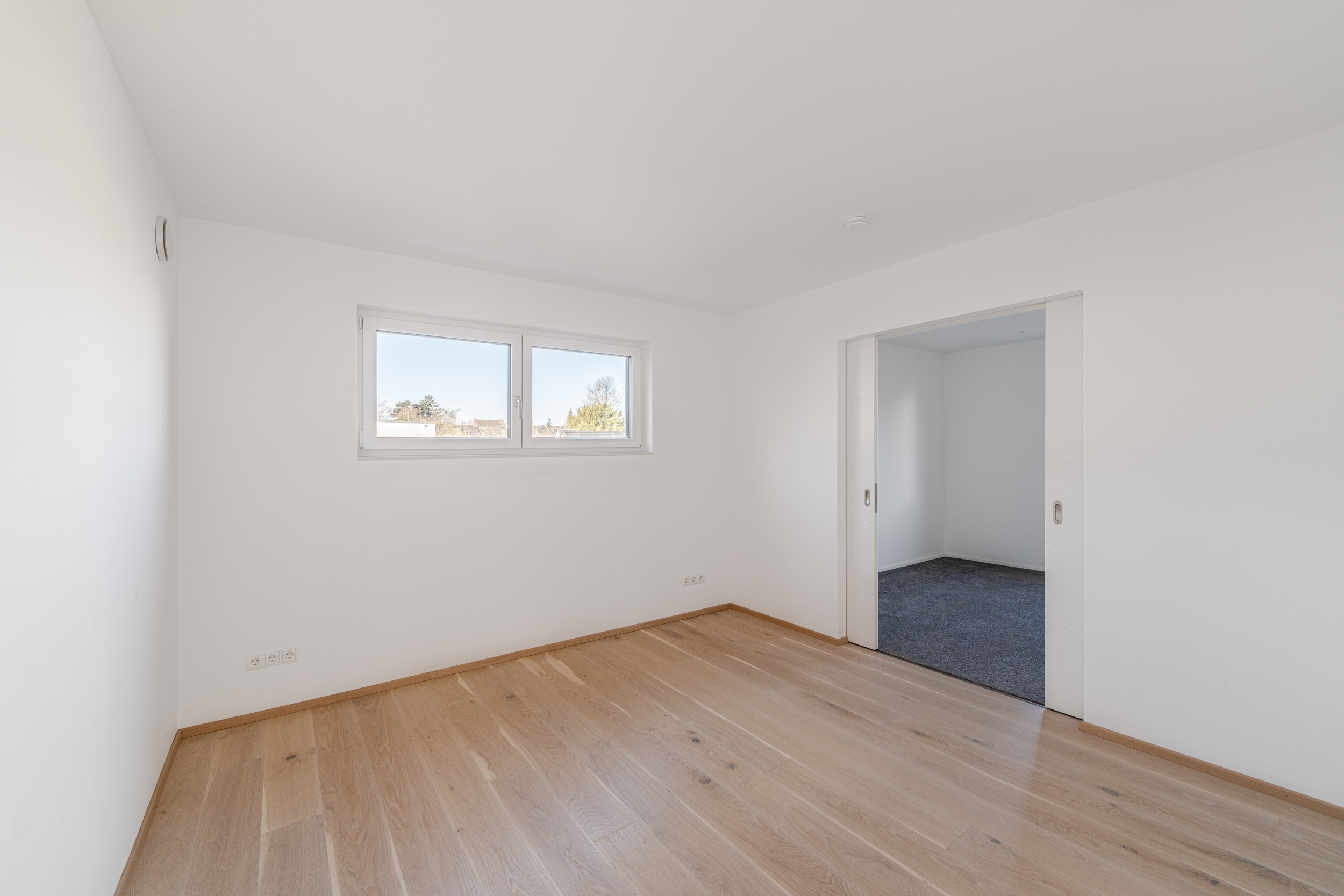 Heller, leerer Raum mit Holzfußboden, zwei Fenstern und Schiebetür zu einem weiteren Zimmer.