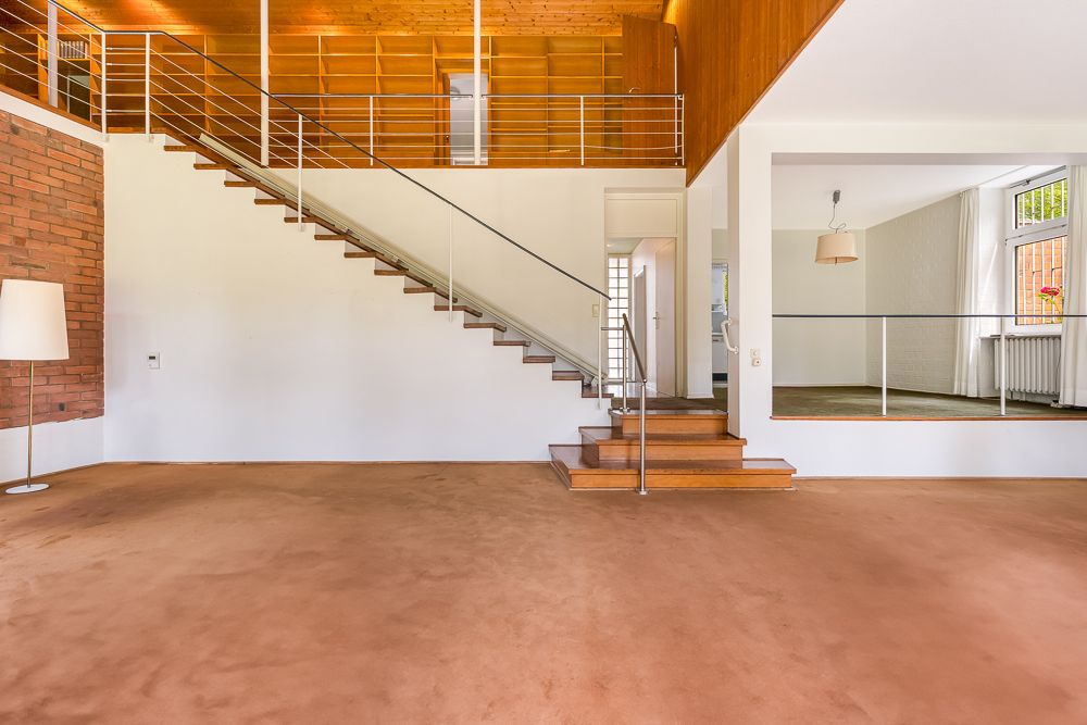Wohnzimmer mit braunem Teppich, Treppe und Blick in ein helles Nebenzimmer.