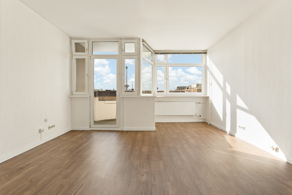 Heller, leerer Raum mit großen Fenstern, weiß gestrichenen Wänden und Holzboden.