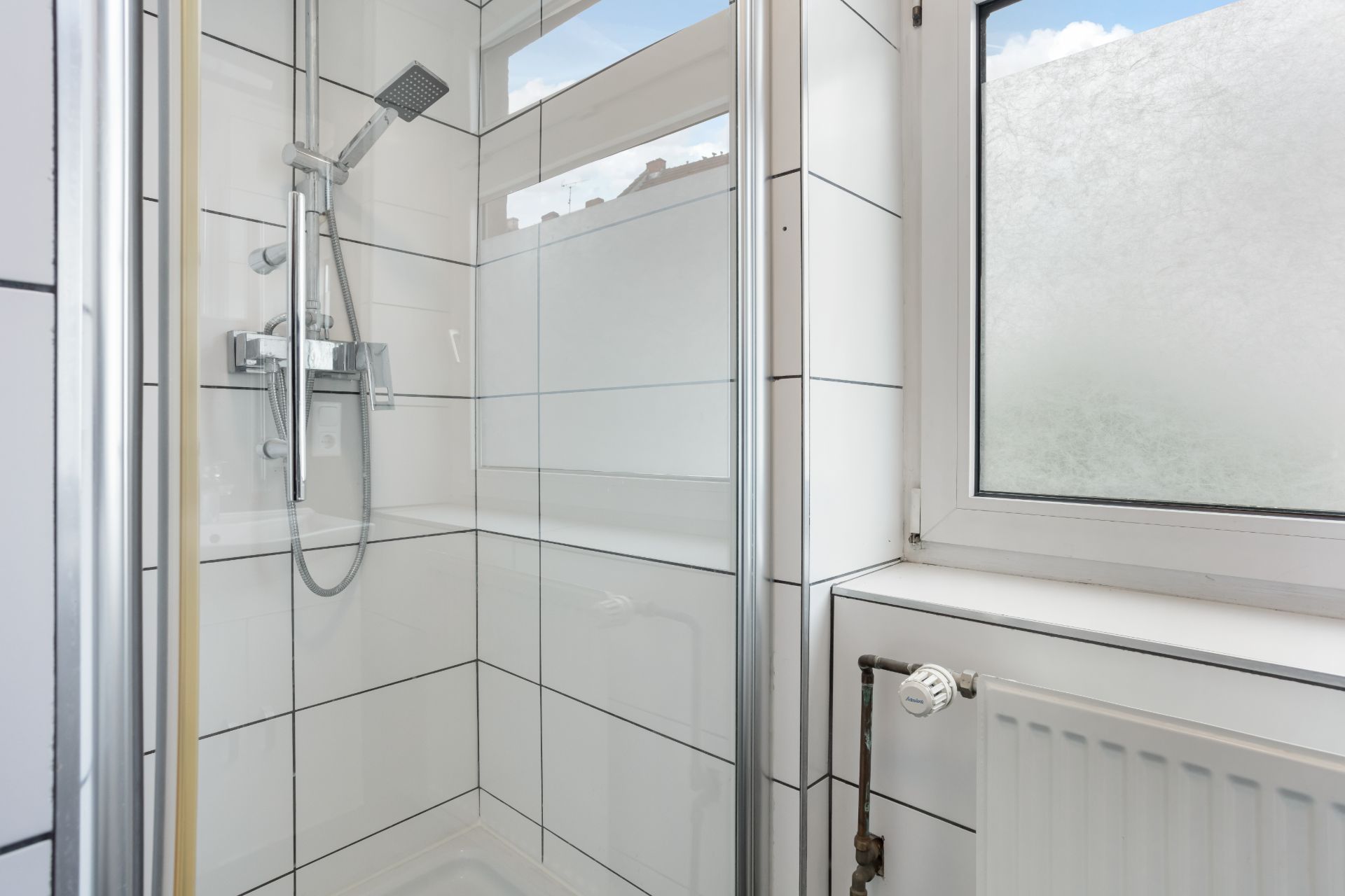 Moderne Dusche mit weiß gefliesten Wänden neben einem Fenster und einem Heizkörper.