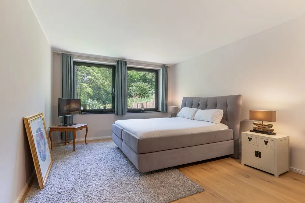 Helles Schlafzimmer mit großem Bett, Teppich, Nachttisch und zwei Fenstern mit Blick ins Grüne.