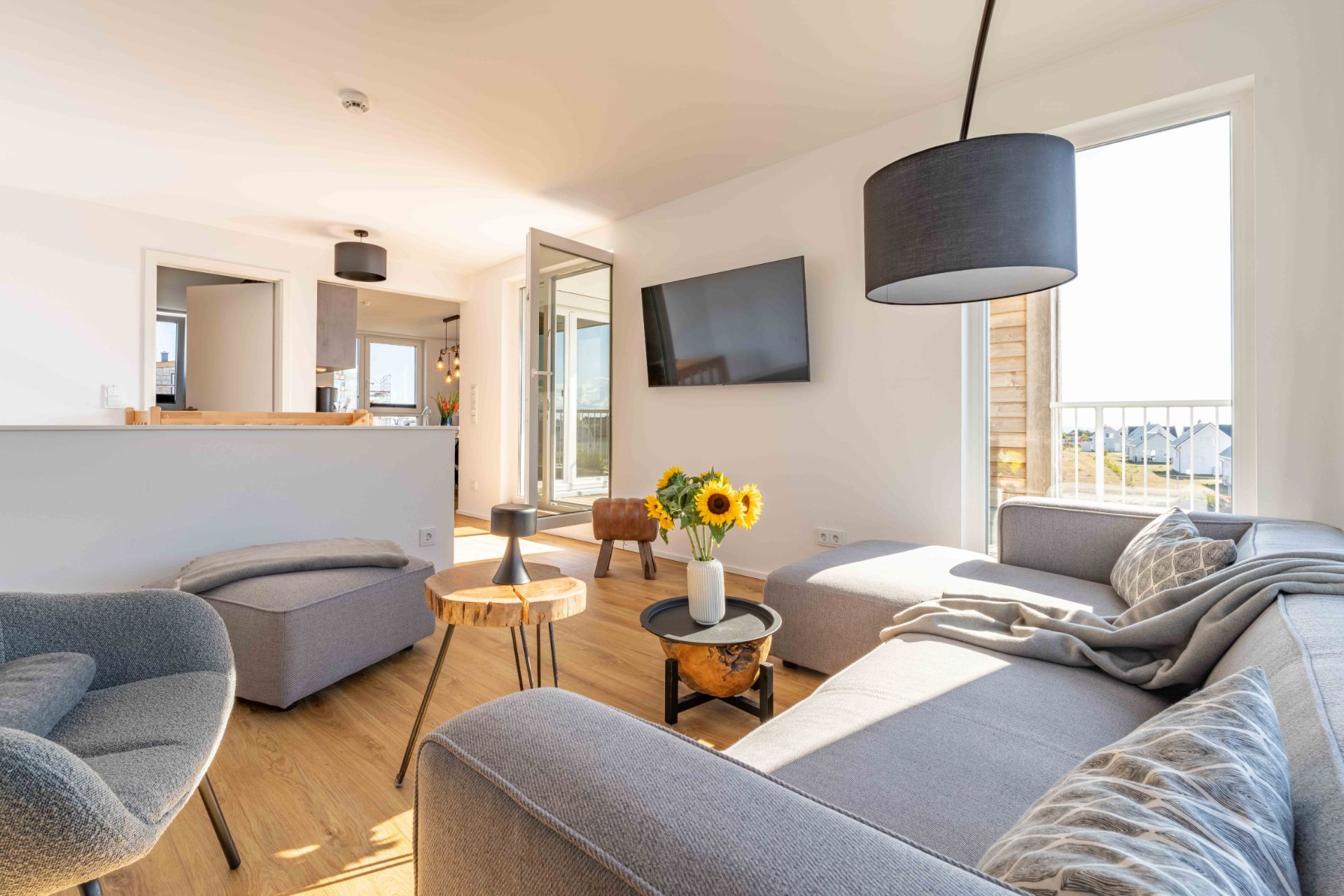 Helles Wohnzimmer mit grauem Sofa, Sessel, Couchtisch, Fernseher und großen Fenstern.